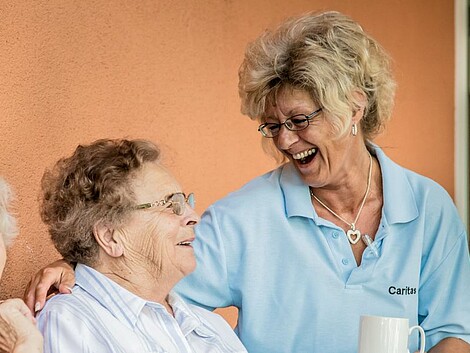 Eine Betreuerin der Caritas und zwei Seniorinnen sitzen lachend auf einer Terrasse. 