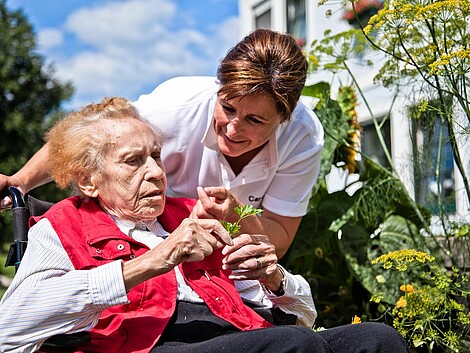 Eine Caritas-Mitarbeiterin spricht mit einer Seniorin im Rollstuhl, die eine Blume in der Hand hält.